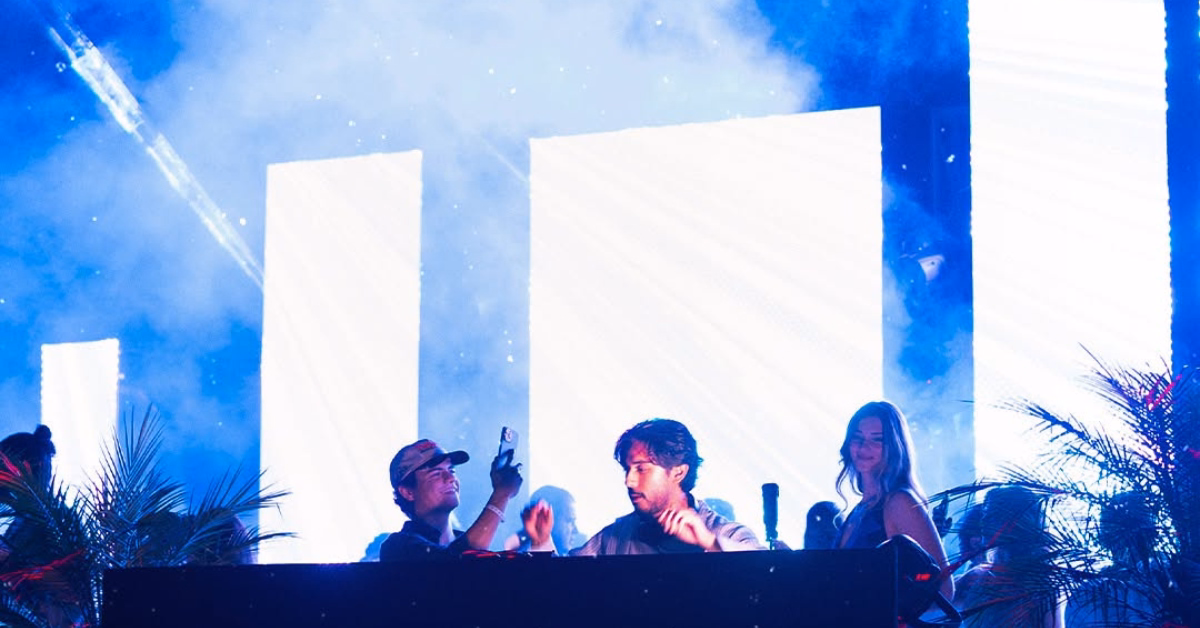 Three people behind a DJ booth at d’cave nightclub, lit by blue and white lights with smoke, enjoying lively music.
