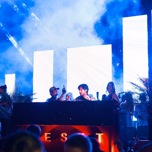 A DJ performs at a booth with vivid LED screens, colorful lights, and smoke as people dance in a lively nightclub atmosphere.