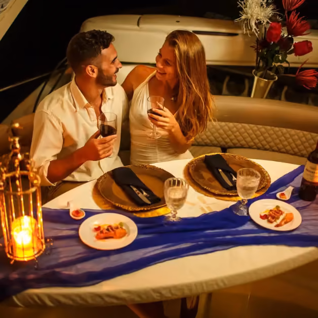 A couple sits close, smiling and sharing wine and appetizers at a candlelit table with flowers and soft romantic lighting.