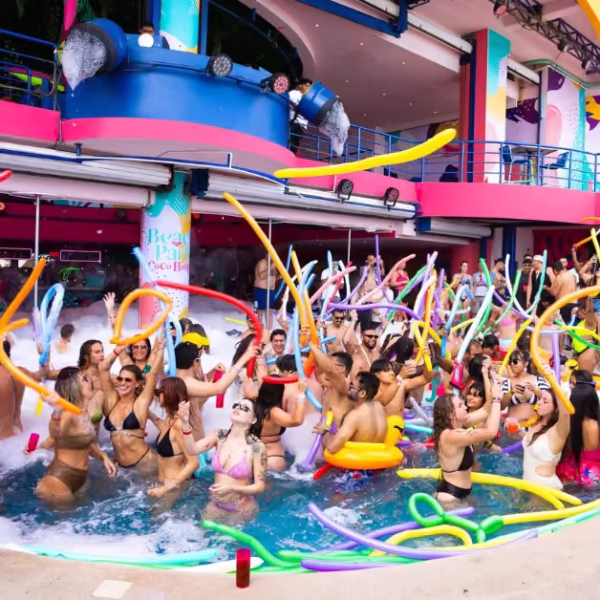 People in swimwear dance and wave colorful foam noodles at a lively city pool party, surrounded by bright decorations.