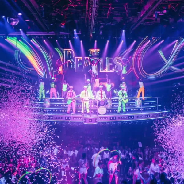 Performers in colorful costumes dance under a neon Beatles sign as confetti falls on a cheering crowd lit by purple lights.