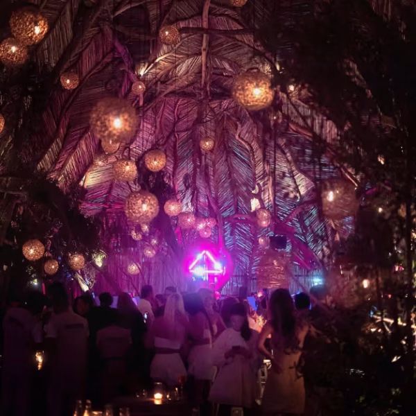 People dance indoors beneath glowing lanterns and a palm ceiling, bathed in soft pink and purple lights at a festive party.