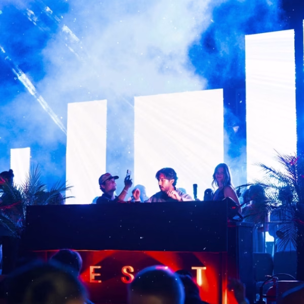 A DJ performs at a booth with vivid LED screens, colorful lights, and smoke as people dance in a lively nightclub atmosphere.