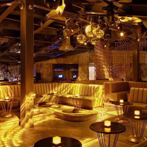 Chic lounge with gold lighting, plush booths, round tables with candles, mirrored disco balls, and a sleek modern vibe.