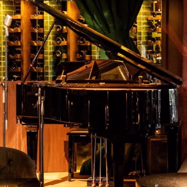 A grand piano sits by a wall of wine bottles and green tiles, with a vintage mic and elegant green curtains above.
