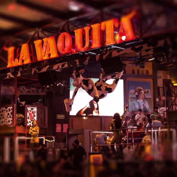 Colorful Cancun nightclub with a neon LA VAQUITA sign, cow-themed decor, socializing guests, and a big cow statue overhead.
