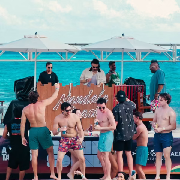 People in swimsuits dance, mingle, and enjoy drinks at a lively beach party by the ocean as a DJ plays music under umbrellas.