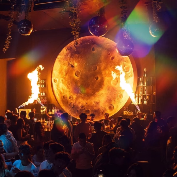 A lively nightclub with a glowing moon backdrop, fire effects, disco balls, and colorful lights shining over dancing partygoers.