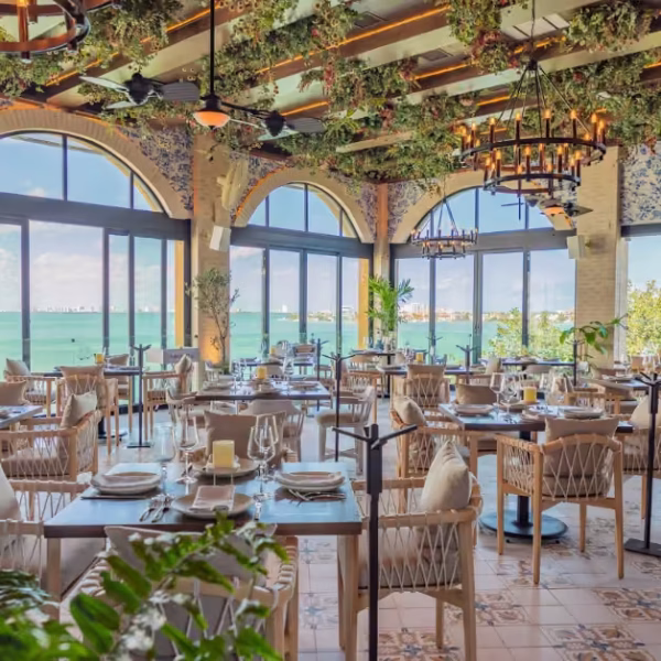 Elegant restaurant with arched windows facing the ocean, hanging plants, chandeliers, and neatly set tables with glassware.