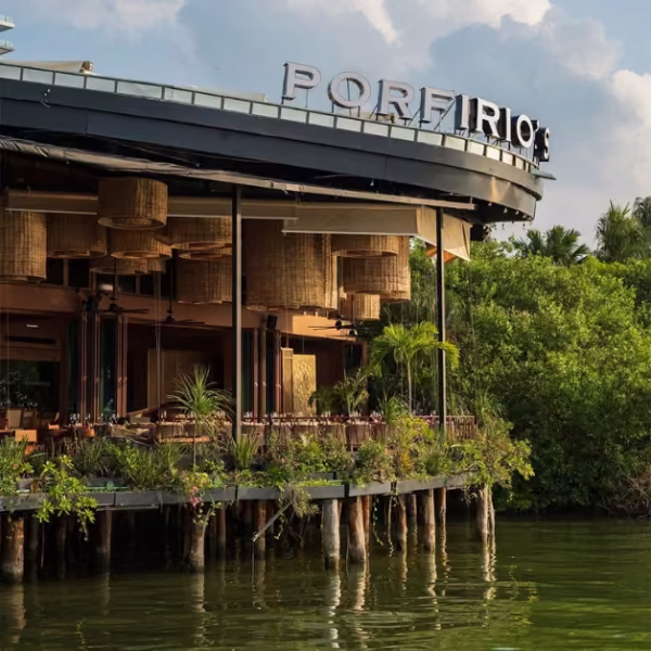 Porfirio’s sits on stilts above the water, surrounded by greenery and hanging lights, with tables under a partly cloudy sky.