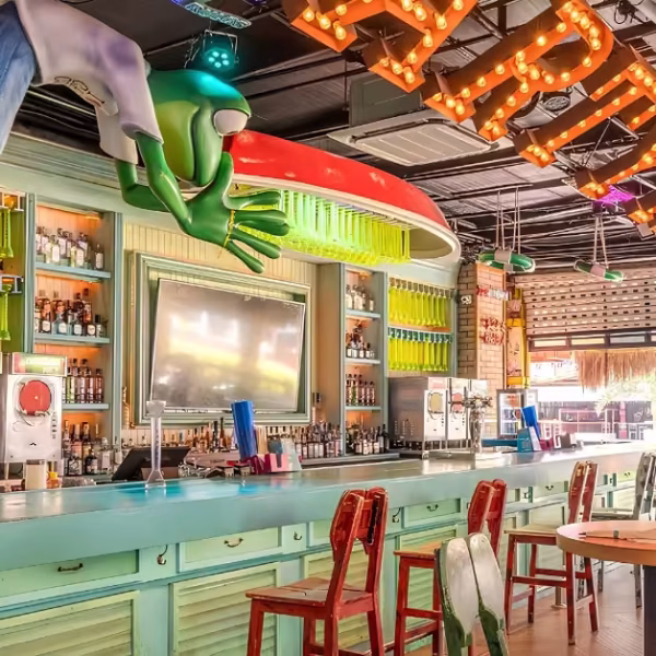 Vibrant bar with pastel counters, red and green chairs, frog decor, "BAR" sign, shelves of bottles, and sunlight through windows.