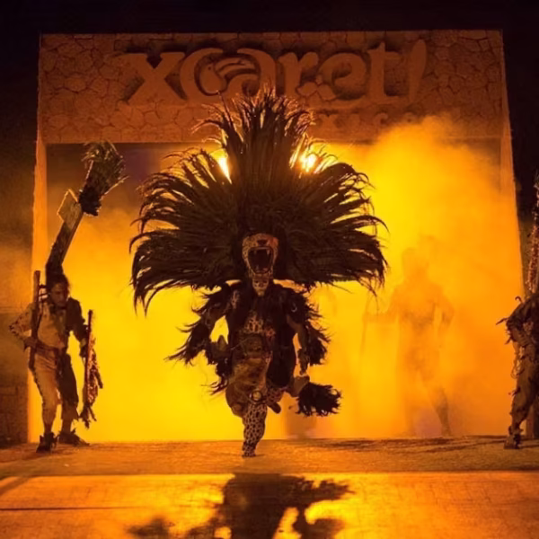 Costumed performers, one in a large feathered headdress, emerge from smoky lights onstage during Xcaret’s vibrant night show.