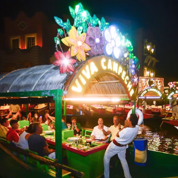 A lively boat with flowers and a Viva Chaparrita sign carries diners along a lit canal at night, with more boats behind.