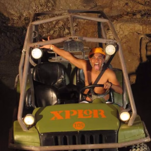 Helmeted driver steers an Xplor off-road vehicle through a rocky cave, arm raised and tongue out as headlights light the walls.