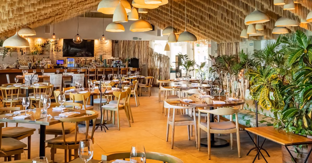 Fred’s Restaurant interior with wooden tables, pendant lights, lush green plants, and a welcoming bar in the background.