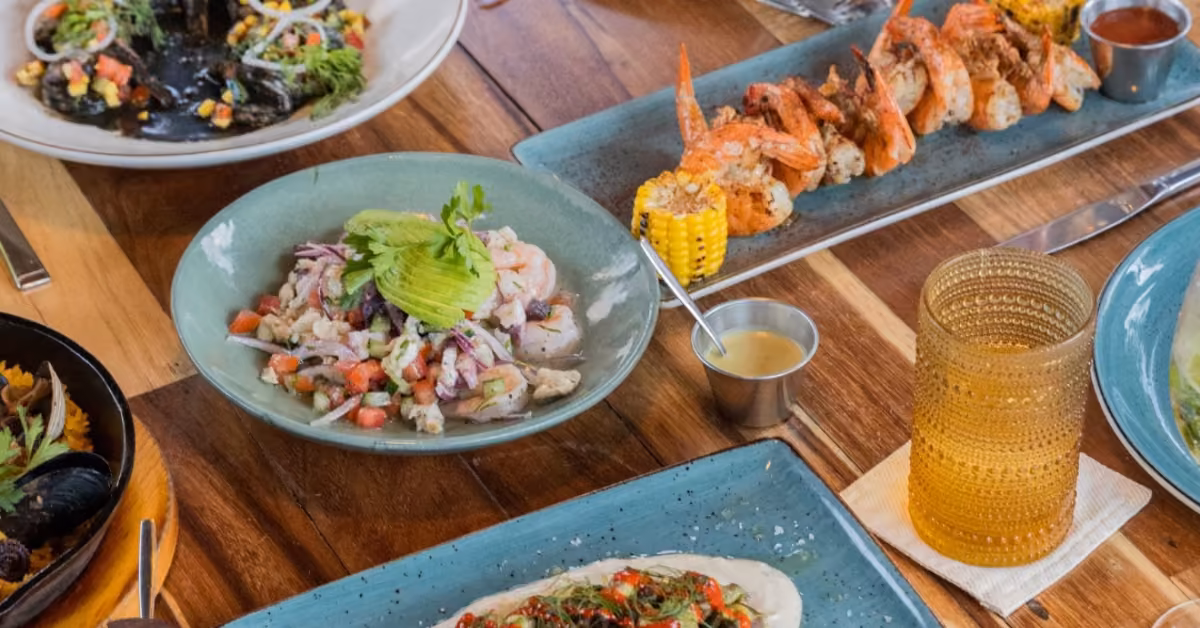 Seafood dishes—shrimp skewers, ceviche with avocado, mussels—served on a wooden table with a yellow glass of water.