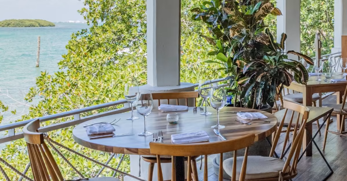 A round table for four overlooks greenery and water on Fred’s patio, set with wine glasses and folded napkins.