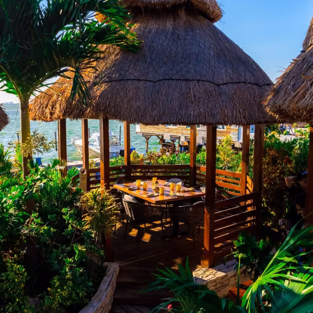Thatched-roof outdoor dining area with lush plants, waterfront view, and a wooden table set for a meal with surrounding chairs.