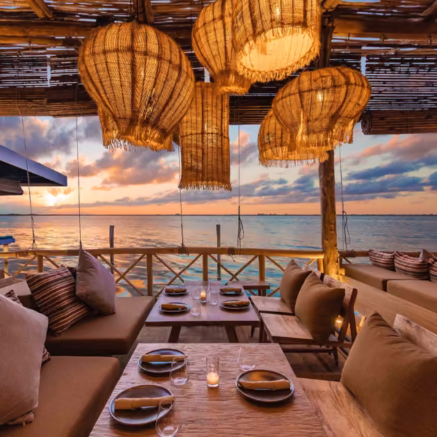 Cozy outdoor restaurant with wooden tables, cushioned seats, woven lamps, and warm lighting overlooking the sea at sunset.