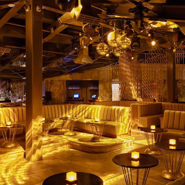 Chic lounge with gold lighting, plush booths, round tables with candles, mirrored disco balls, and a sleek modern vibe.