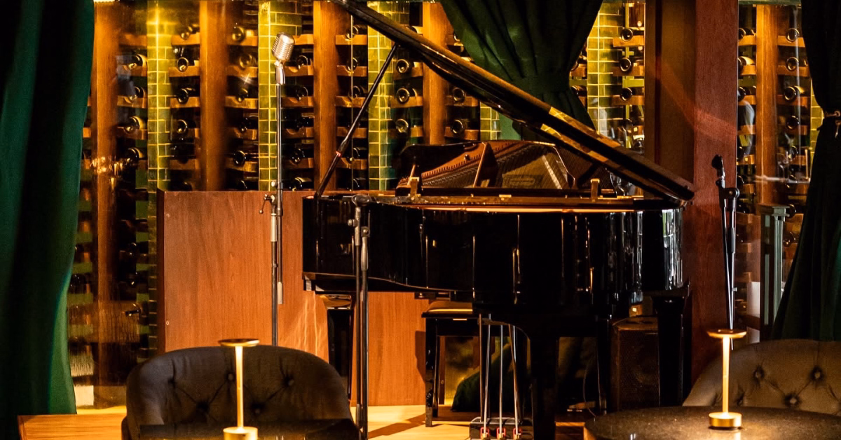 A grand piano and vintage microphone on a small stage with green curtains and wine bottles create an elegant, intimate vibe.