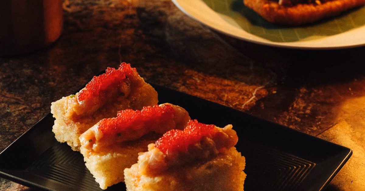 Three sushi pieces with red roe on a black plate, atop a dark marbled table. A white plate is blurred in the background.