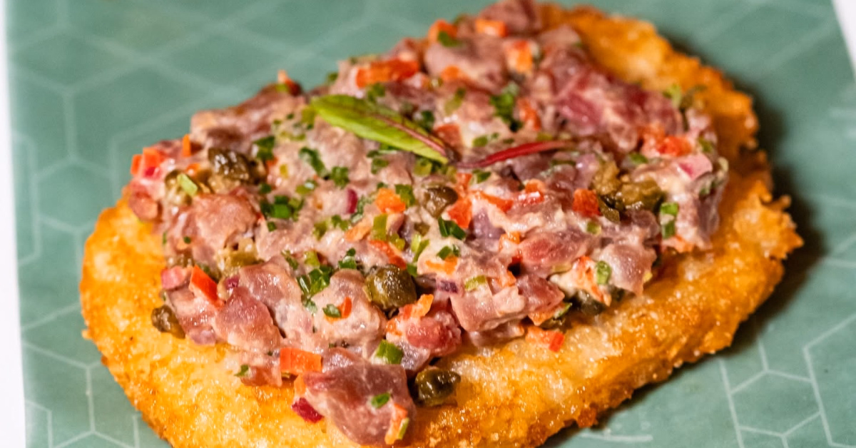Golden hash brown topped with diced raw tuna, capers, red onion, and herbs on a green geometric-patterned plate.