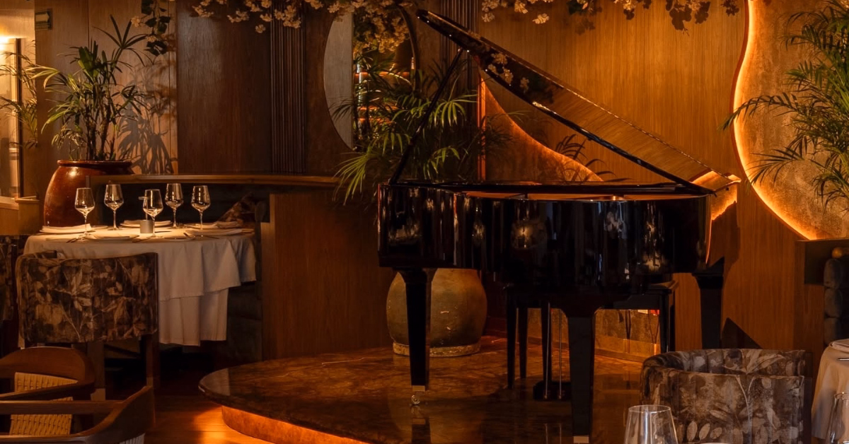 A black grand piano on a small stage in an elegant restaurant amid plants and white-clothed tables with warm, cozy lighting.