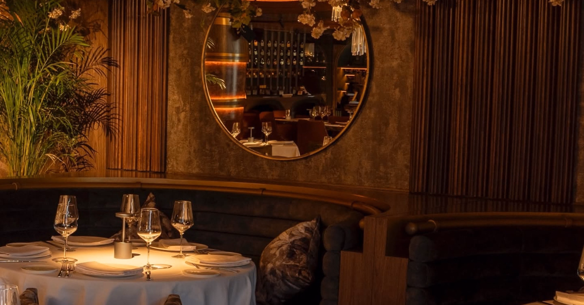 A cozy, dim restaurant with a round table for four, wine glasses, a candle, mirror reflections, curved seats, and plants.