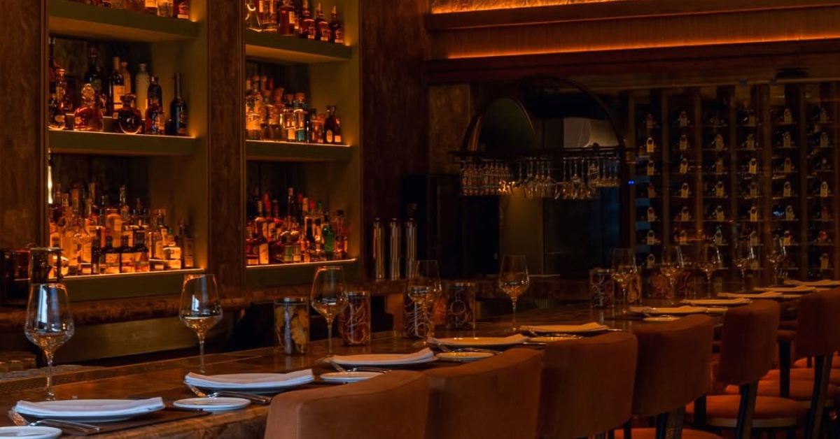Harry’s Bar, softly lit and elegant, has shelves of liquor, a wooden counter with cushioned chairs, and tables with wine glasses.