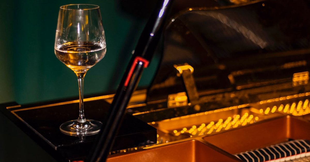 A wine glass with white wine rests on a glossy black piano, its strings and gold interior softly visible in the background.