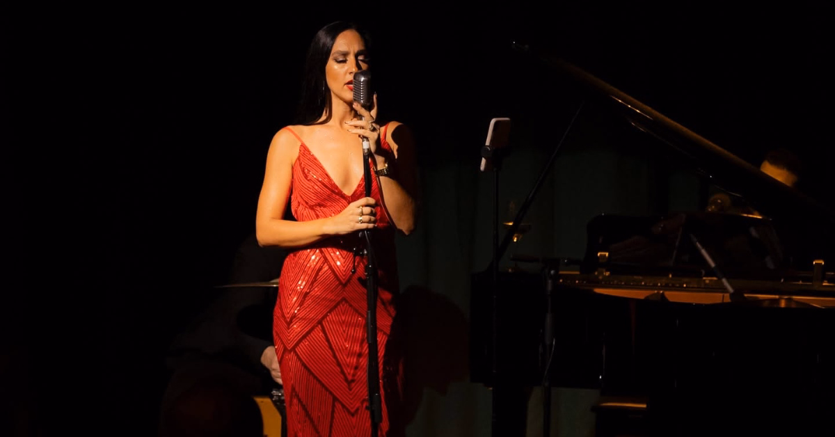 A woman in a red dress sings into a vintage mic on a dim stage at Harry’s Bar, with a piano and instruments behind her.