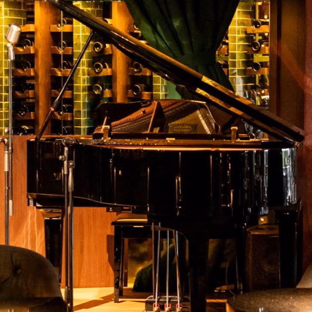 A grand piano sits by a wall of wine bottles and green tiles, with a vintage mic and elegant green curtains above.