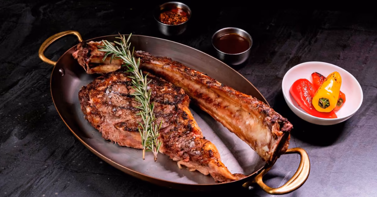 Tomahawk steak with rosemary in an oval dish at Harry’s, served with grilled mini bell peppers and sauces on a dark table.