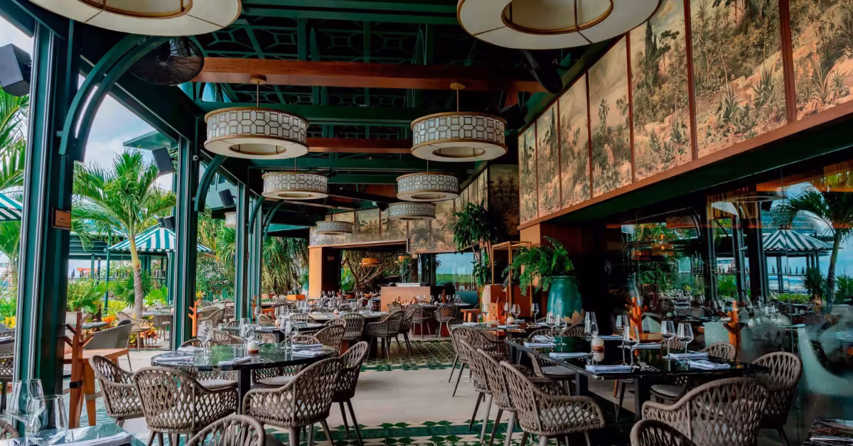 Harry’s Restaurant offers wicker chairs, set tables, lush plants, and round lights under striped canopies with greenery outside.