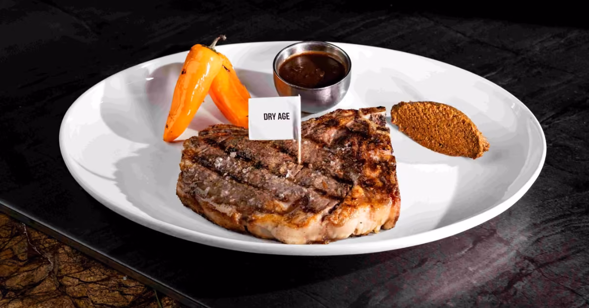 A grilled steak at Harry’s Restaurant on a white plate with roasted orange pepper, brown seasoning, and a small cup of sauce.