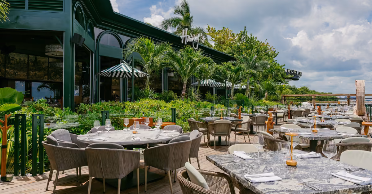 Patio at Harry’s Restaurant with marble tables, arranged chairs, striped umbrellas, lush greenery, and palm trees outdoors.
