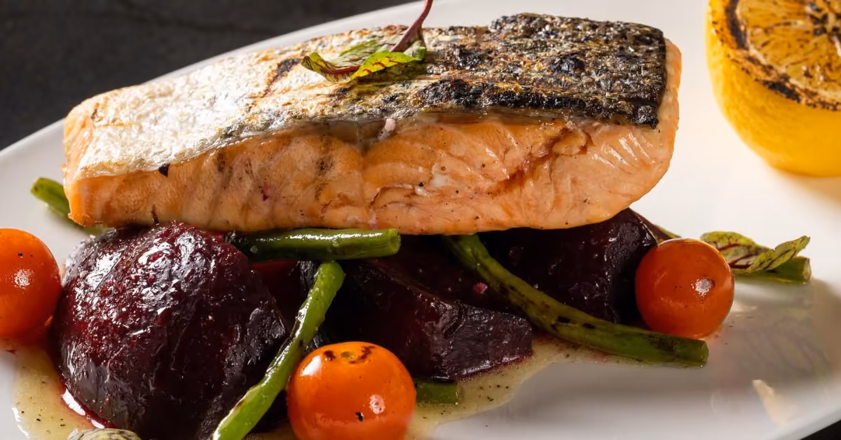 Crispy salmon fillet on roasted beets, green beans, and cherry tomatoes, topped with herbs and grilled lemon on a white plate.