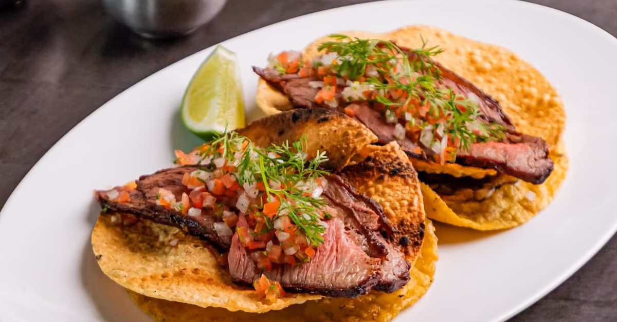 Two grilled steak tacos with tomatoes, onions, herbs, and greens on crispy tortillas, served with a lime wedge.