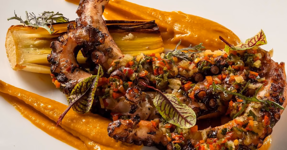 Grilled octopus tentacle topped with diced veggies and herbs on orange sauce, served with roasted leeks on a white plate.