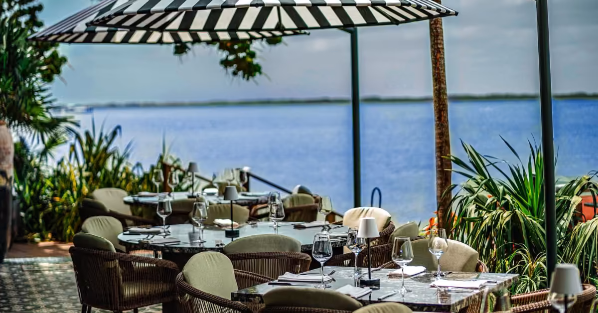 Harry’s patio with wicker chairs and tables beneath a striped umbrella, overlooking calm water and lush greenery.