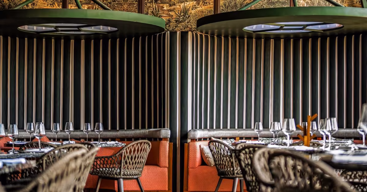 Harry’s cozy interior with woven chairs, orange cushions, set tables with wine glasses, and round lights against paneled walls.