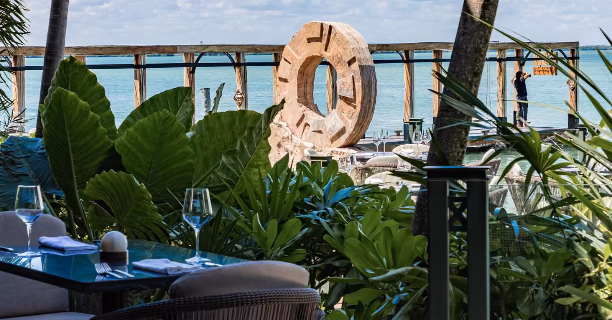Harry’s Restaurant has a glass table and chairs by the ocean, surrounded by greenery and a stone sculpture near a wooden pier.