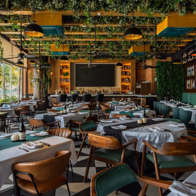 Modern restaurant with wood chairs, white tablecloths, checkered floor, hanging greenery, warm lights, and large windows.
