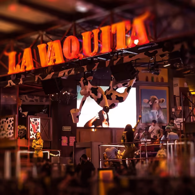 Colorful Cancun nightclub with a neon LA VAQUITA sign, cow-themed decor, socializing guests, and a big cow statue overhead.