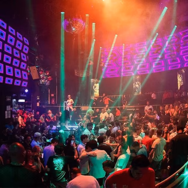 A lively nightclub with colorful lights, people dancing and chatting, and glowing geometric patterns on the walls and ceiling.