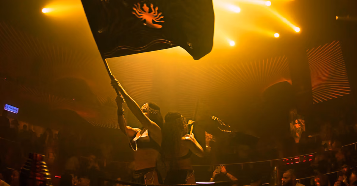 Two people wave black flags with sun symbols under yellow stage lights in a lively, packed nightclub with dramatic lighting.