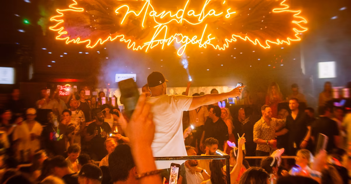A vibrant nightclub with a DJ on stage, neon Mandalas Angels sign overhead, and a cheering crowd holding up phones and drinks.