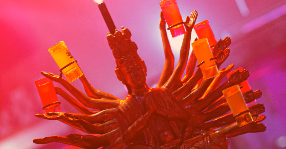 A multi-armed statue holding colorful drinks under pink and red lights at Mandala Night Club, with a candle on its head.