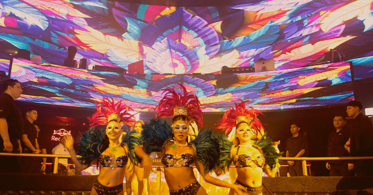 Three showgirls in colorful costumes and feathered headdresses pose on stage under bright lights at Mandala Night Club.